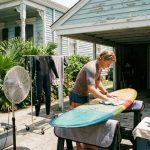 Surfer drying surfboard and wetsuit in shaded outdoor area during humid summer weather in New Orleans Louisiana