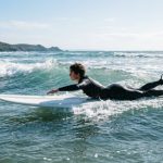 A surfer wearing a black wetsuit paddling out through ocean waves on a sunny day