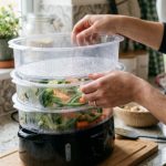 A modern electric food steamer filled with freshly steamed vegetables on a kitchen counter.