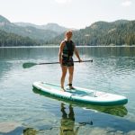 Beginner woman balancing on an inflatable paddle board on a calm lake.